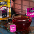 A glass teacup filled with dark berry tea sits on a matching saucer, with a pink Miles Berry tag hanging from the handle. A yellow and pink Miles Berry tea box is visible in the background along with a pink teapot and a blurred tea bag.