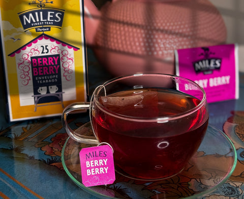 A glass teacup filled with dark berry tea sits on a matching saucer, with a pink Miles Berry tag hanging from the handle. A yellow and pink Miles Berry tea box is visible in the background along with a pink teapot and a blurred tea bag.