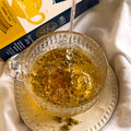 A glass teacup filled with Camomile tea, with hot water being poured into it. A yellow and navy herbal tea box labeled 'Camomile Flower' is visible in the background on draped white fabric.
