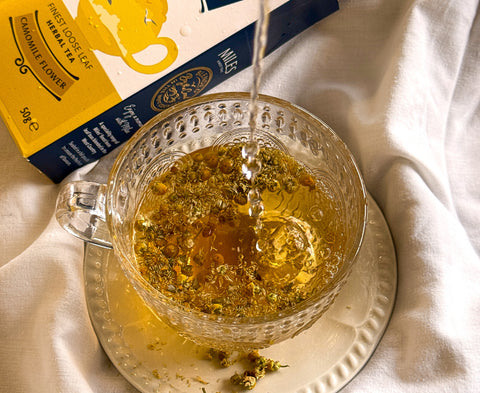 A glass teacup filled with Camomile tea, with hot water being poured into it. A yellow and navy herbal tea box labeled 'Camomile Flower' is visible in the background on draped white fabric.