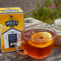 A glass teacup filled with amber Earl Grey tea and a lemon slice sits on a wooden table beside a branded Miles of London tea box and a teapot, with an outdoor garden blurred in the background.