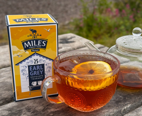A glass teacup filled with amber Earl Grey tea and a lemon slice sits on a wooden table beside a branded Miles of London tea box and a teapot, with an outdoor garden blurred in the background.