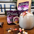 A styled product shot of Miles Homemade Hot Chocolate, showing a ceramic mug filled with whipped cream, chocolate drizzle, and marshmallows, alongside a canister and a paper bag of the hot chocolate mix on a rustic wooden surface.