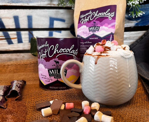 A styled product shot of Miles Homemade Hot Chocolate, showing a ceramic mug filled with whipped cream, chocolate drizzle, and marshmallows, alongside a canister and a paper bag of the hot chocolate mix on a rustic wooden surface.