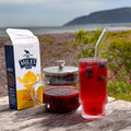 A box of Miles Lemon & Mango tea sits on a weathered wooden table outdoors, next to a glass teapot filled with dark tea and a tall glass of red tea with blueberries and a glass straw. The background shows a blurred coastal landscape with sea, hills, and greenery.