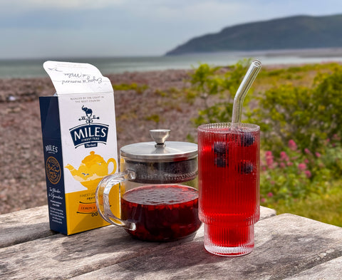 A box of Miles Lemon & Mango tea sits on a weathered wooden table outdoors, next to a glass teapot filled with dark tea and a tall glass of red tea with blueberries and a glass straw. The background shows a blurred coastal landscape with sea, hills, and greenery.