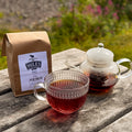 A brown paper bag of Miles Nilgiri Tea sits on a weathered wooden table next to a clear glass teapot and a matching glass teacup filled with the dark tea, with a blurred garden background.