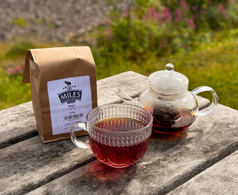 A brown paper bag of Miles Nilgiri Tea sits on a weathered wooden table next to a clear glass teapot and a matching glass teacup filled with the dark tea, with a blurred garden background.