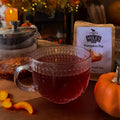 Tea cup with red liquid, pumpkin spice tea package, and small pumpkin on a wooden surface.