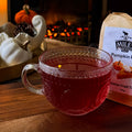 Glass mug with red liquid next to a package of Miles Pumpkin Pie tea on a wooden surface.