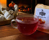 Glass mug with red liquid next to a package of Miles Pumpkin Pie tea on a wooden surface.
