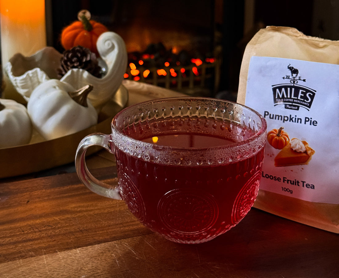 Glass mug with red liquid next to a package of Miles Pumpkin Pie tea on a wooden surface.