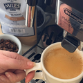 A close-up lifestyle shot of a person's hand holding a white ceramic mug under the hot water spout of a De'Longhi Magnifica Evo espresso machine, with a bag of Miles Coffee Roasters Kenya Roast coffee beans and a small bowl of roasted coffee beans in the background.