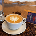 A cup of latte coffee with heart-shaped latte art sits on a saucer, surrounded by coffee beans, with a MILES brand coffee bean bag package and part of a moka pot visible in a styled coffee setting.