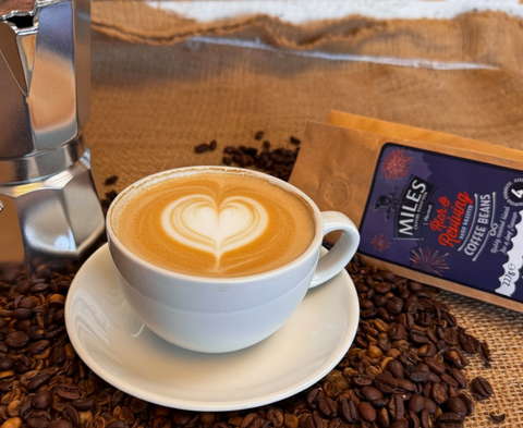 A cup of latte coffee with heart-shaped latte art sits on a saucer, surrounded by coffee beans, with a MILES brand coffee bean bag package and part of a moka pot visible in a styled coffee setting.