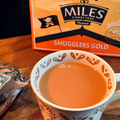 A cup of tea on a wooden table with a box of Miles Finest Teas Smugglers Gold tea blend in the background.