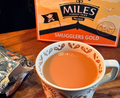 A cup of tea on a wooden table with a box of Miles Finest Teas Smugglers Gold tea blend in the background.