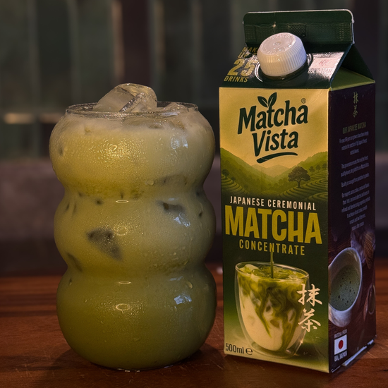 Matcha Vista matcha concentrate box next to a green drink with ice cubes on a wooden surface.