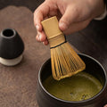 Person using a wooden whisk to stir matcha in a black cup on a wooden surface.