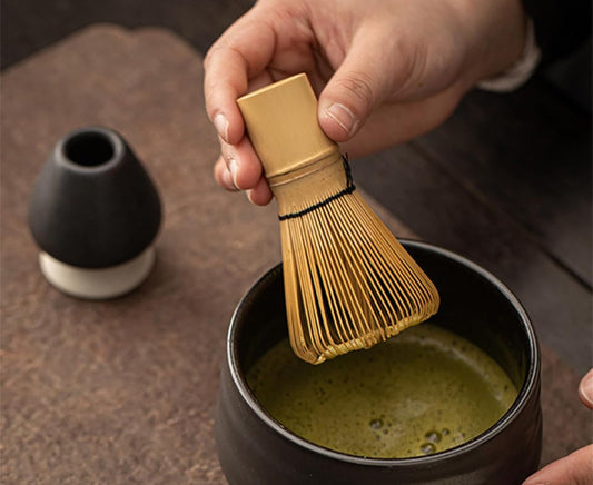 Person using a wooden whisk to stir matcha in a black cup on a wooden surface.
