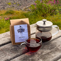 A paper bag of Miles Yunnan tea sits on a weathered wooden table next to a glass teapot filled with tea and a glass cup of the same dark red tea, with a blurred outdoor garden background.