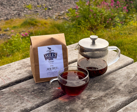 A paper bag of Miles Yunnan tea sits on a weathered wooden table next to a glass teapot filled with tea and a glass cup of the same dark red tea, with a blurred outdoor garden background.